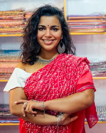 A smiling woman in a red sequinned saree standing in front of shelves of folded sarees